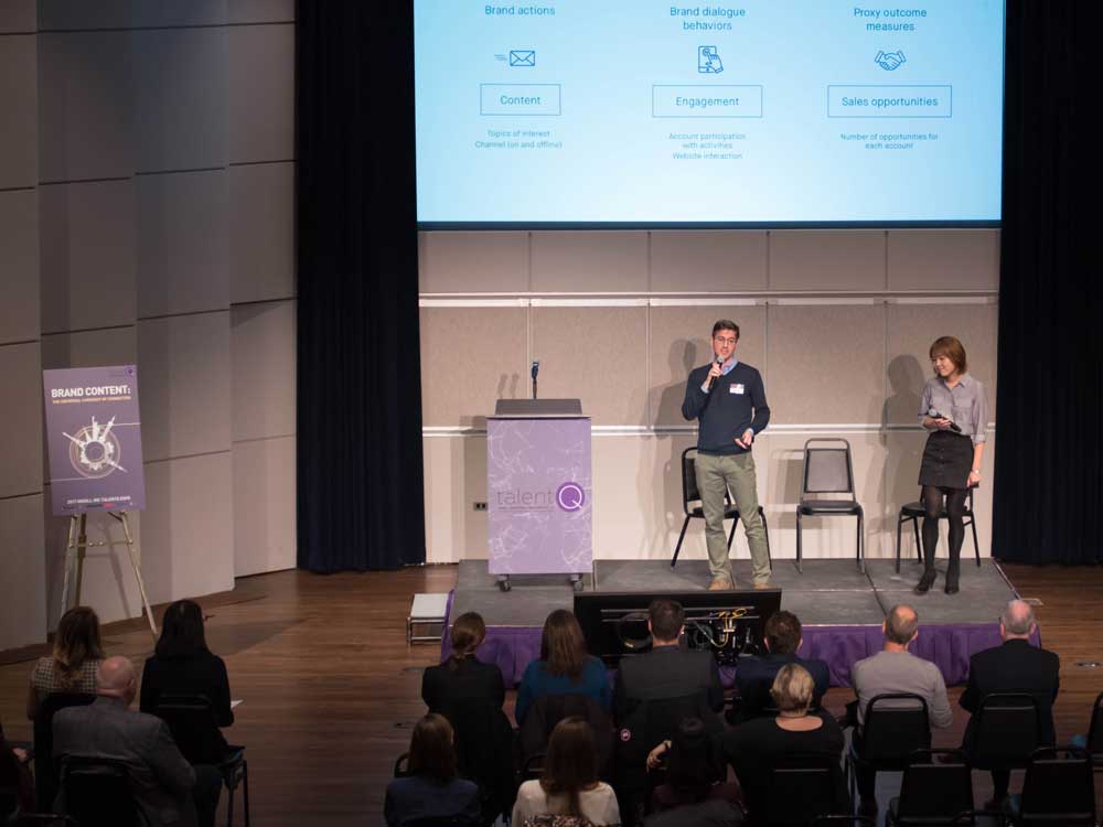 A male student and a female student stand on stage in front of a large screen and speak to the audience