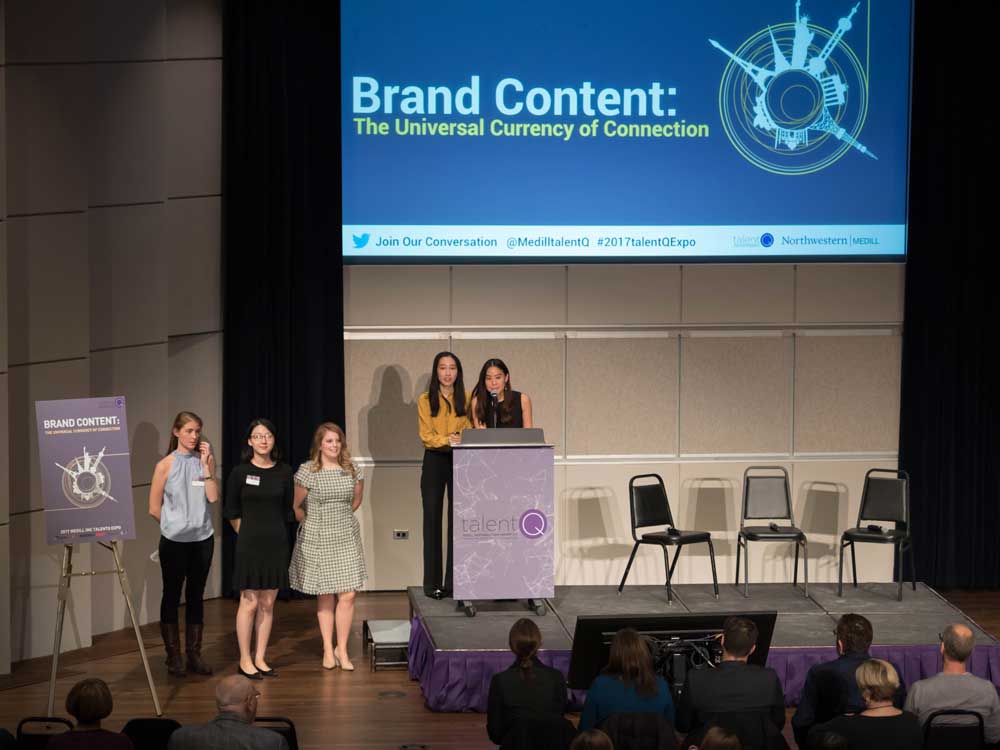 Five students stand at the front of an auditorium; one of them speaks into a microphone at a podium