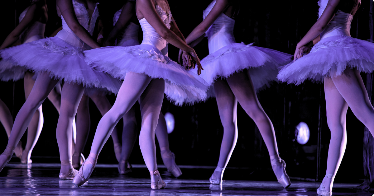 Photo of ballet dancers on stage from the waist down in a purple hue with stage lights shining on them.