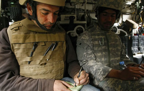 Two men in military gear sit inside a vehicle. The man on the left writes in a notebook, appearing focused. The man on the right observes quietly.